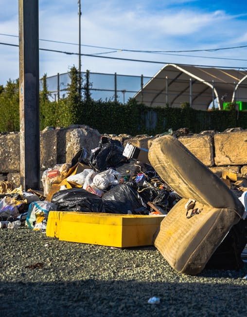 A roadside collection of assorted waste and rubbish situated against a low stone wall, with a variety of black plastic rubbish bags, cardboard boxes, and loose debris visible in the scene. Prominently featured are a large, old, beige car tire leaning on its side in the foreground, partially obscuring smaller waste items around it. The ground appears to be gravel or asphalt, with some scattered litter. Behind the waste, there is a backdrop of green hedges and trees, along with a modern sports or tennis court fence, and a large curved canopy structure under a bright, partly cloudy blue sky. The image conveys an informal bulk waste collection scenario, likely associated with private disposal services like Rubbish Removal Surrey, illustrating the type of clutter that might require professional rubbish clearance or alternative waste handling solutions alongside or independent from local authority waste collection.