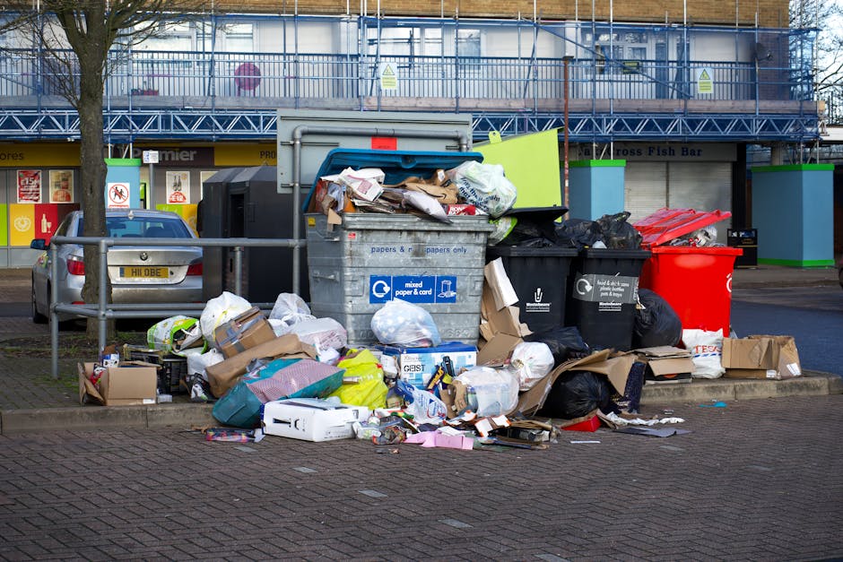 A large pile of mixed household and commercial waste, including cardboard boxes, plastic bags, paper, and discarded packaging, is overflowing from black and gray rubbish bins situated on a paved urban street. The bins are positioned near a metal railing and directly in front of a building with shuttered storefronts and a blue scaffolding structure, indicating ongoing maintenance or renovation. The waste extends onto the sidewalk and partially onto the road, with some items spilling onto the street surface. The scene is illuminated by natural daylight, with a tree on the left side and parked cars nearby, highlighting the importance of careful waste management and potential private disposal options. The cluttered arrangement suggests improper disposal or overfilled bins, which could relate to issues surrounding alternative waste handling methods that bypass local authority collection. Rubbish Removal Surrey specializes in rubbish services, making this scene relevant to discussions about avoiding hidden charges through independent collection or on-site clearance solutions.