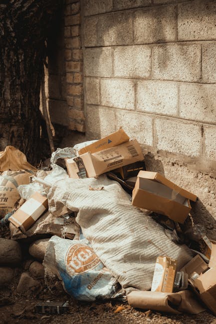 A large pile of mixed household and commercial waste, including cardboard boxes, plastic bags, paper, and discarded packaging, is overflowing from black and gray rubbish bins situated on a paved urban street. The bins are positioned near a metal railing and directly in front of a building with shuttered storefronts and a blue scaffolding structure, indicating ongoing maintenance or renovation. The waste extends onto the sidewalk and partially onto the road, with some items spilling onto the street surface. The scene is illuminated by natural daylight, with a tree on the left side and parked cars nearby, highlighting the importance of careful waste management and potential private disposal options. The cluttered arrangement suggests improper disposal or overfilled bins, which could relate to issues surrounding alternative waste handling methods that bypass local authority collection. Rubbish Removal Surrey specializes in rubbish services, making this scene relevant to discussions about avoiding hidden charges through independent collection or on-site clearance solutions.