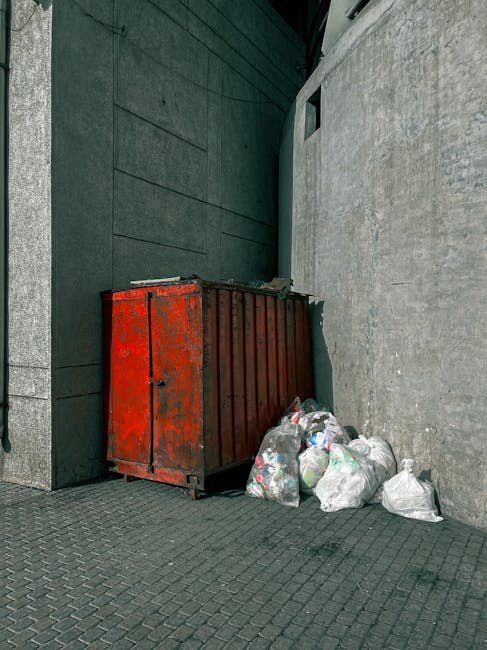 A large, rusted red metal waste container with vertical ridges sits against a dark green brick wall and a light grey concrete wall on a paved outdoor surface. Nearby, several plastic bags filled with mixed household rubbish are piled close to the container, some slightly torn or bulging. The scene is set in a narrow urban alleyway or corner, with shadows indicating natural daylight. The container appears used for on-site waste collection, typical of private rubbish removal services. The environment external to a commercial or residential building suggests an area where independent rubbish disposal occurs, supporting local waste removal options outside of regular council services. The ground consists of small, interlocking dark grey paving stones, and the overall setting remains utilitarian and functional, focusing on waste storage and collection without decorative elements.