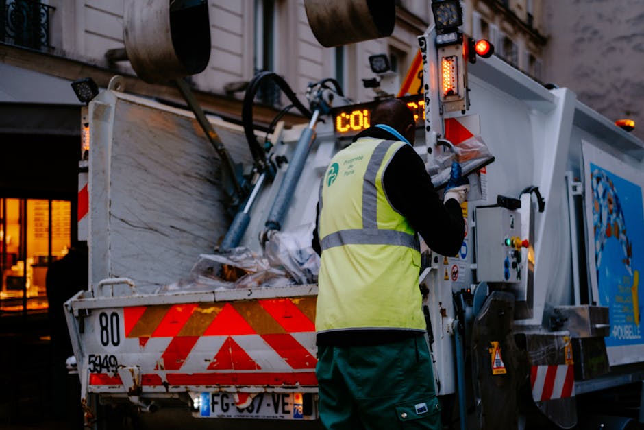 A waste management worker wearing a high-visibility yellow safety vest and dark clothing is operating controls on a large, modern rubbish collection vehicle parked on a city street. The vehicle features a white metal body with red and orange reflective warning markings along its edges for safety. The rear section of the truck is open, revealing collected waste, primarily consisting of mixed rubbish such as crumpled paper, plastic, and other discarded materials. Visible on the vehicle are various hydraulic and control components with digital displays, along with warning lights illuminated in red and amber. The background includes parts of urban buildings illuminated by warm indoor lighting, with an ambient evening atmosphere suggested by the limited natural light. The worker appears to be managing on-site waste collection or disposal, aligning with services provided by companies like Rubbish Removal Surrey for private waste handling or alternative rubbish collection, particularly suited for tight access jobs or special disposal requirements. The scene emphasizes professional waste collection processes conducted in urban environments, supporting the context of independent rubbish removal services.