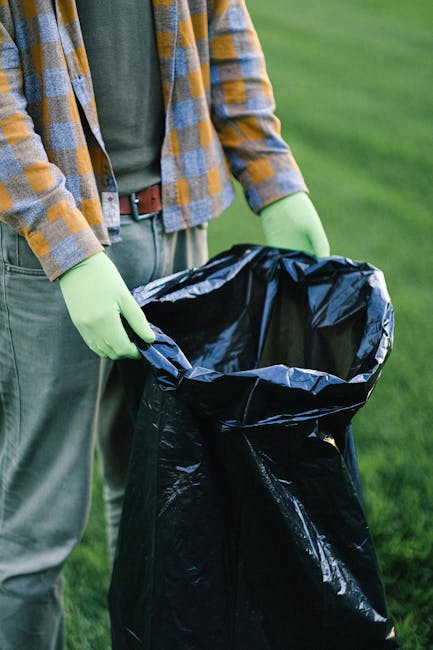 Close-up view of a person wearing a green long-sleeved shirt, a yellow and gray plaid jacket, and light green rubber gloves, standing outdoors on a grassy area. They are holding open a large black trash bag, which appears to be made of plastic with a shiny finish, ready for waste disposal. The grassy environment in the background suggests a garden or outdoor space, indicating at-home or garden waste collection possibly coordinated by a service such as Rubbish Removal Surrey. The person's hands are positioned at the top of the open bag, prepared for rubbish collection, highlighting an active waste clearance process. The scene emphasizes the practical aspects of rubbish handling, consistent with private or alternative waste disposal methods away from municipal services, aligning with the context of on-site clearance or independent rubbish removal. The natural lighting illuminates the scene evenly, underscoring a professional and factual approach to waste management tasks in a residential or outdoor setting.
