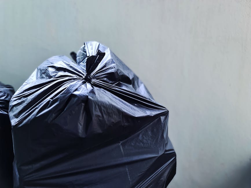 Close-up of multiple black plastic rubbish bags sat against a light-colored, smooth wall. The bags are made from shiny, crinkled plastic material, tightly knotted at the top, with some showing slight bulges indicating they contain waste. They are positioned on the ground, with one bag prominently shown in the foreground and partially leaning on others behind it. The environment appears to be an outdoor or semi-outdoor area, such as a driveway or back alley, with no other objects or debris visible. The lighting is natural and even, emphasizing the reflective surface of the plastic bags. The scene reflects a typical setting for private waste collection or rubbish removal, as offered by Rubbish Removal Surrey, and illustrates an instance of on-site clearance or prepared waste ready for collection.