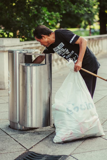 Close-up view of a person wearing a green long-sleeved shirt, a yellow and gray plaid jacket, and light green rubber gloves, standing outdoors on a grassy area. They are holding open a large black trash bag, which appears to be made of plastic with a shiny finish, ready for waste disposal. The grassy environment in the background suggests a garden or outdoor space, indicating at-home or garden waste collection possibly coordinated by a service such as Rubbish Removal Surrey. The person's hands are positioned at the top of the open bag, prepared for rubbish collection, highlighting an active waste clearance process. The scene emphasizes the practical aspects of rubbish handling, consistent with private or alternative waste disposal methods away from municipal services, aligning with the context of on-site clearance or independent rubbish removal. The natural lighting illuminates the scene evenly, underscoring a professional and factual approach to waste management tasks in a residential or outdoor setting.